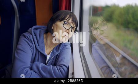 Une femme âgée en verre voyage en train et regarde la fenêtre reflétée dans le verre Banque D'Images