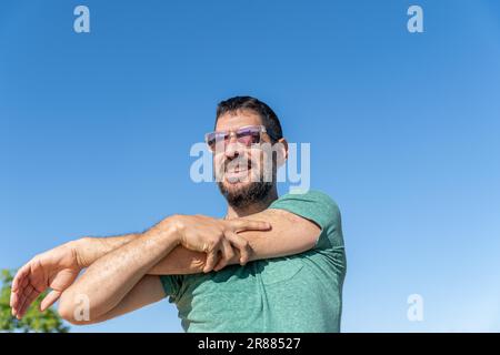 Vue de face d'un bel homme barbu avec des lunettes de soleil souriant et étirant ses muscles dans un champ avec un ciel bleu en arrière-plan Banque D'Images