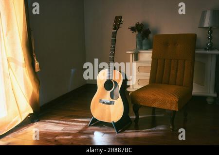 Une guitare acoustique espagnole sur un stand à côté d'une chaise dans l'ombre d'une pièce sombre avec une lumière vive venant de derrière un rideau Banque D'Images