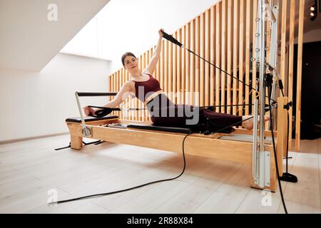 Une jeune fille fait Pilates sur un lit reformeur dans un studio lumineux. Un brunette mince dans un bodysuit bordeaux fait des exercices pour renforcer les bras an Banque D'Images