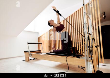 Une jeune fille fait Pilates sur un lit reformeur dans un studio lumineux. Un brunette mince dans un bodysuit bordeaux fait des exercices pour renforcer les bras an Banque D'Images