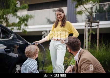 Portriat de la femme qui charge la voiture électrique dans la rue, tandis que mari et fils la regardant. Banque D'Images