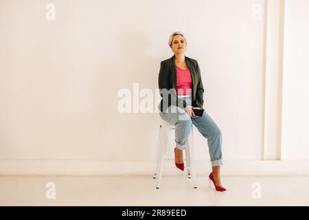 femme d'affaires regardant la caméra tout en étant assise sur une chaise avec un smartphone dans ses mains. Jeune femme d'affaires confiante assise contre un mur dos Banque D'Images