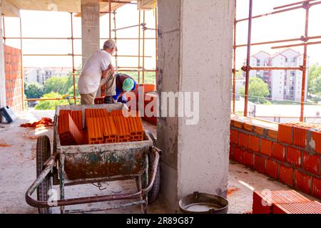 Tas de blocs rouges dans la brouette industrielle obsolète en attente d'être installé, en arrière-plan le mason est la construction d'un mur avec des briques rouges en céramique. Édifice Banque D'Images