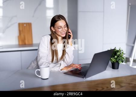 Photo d'une jeune femme d'intérieur à joyeuse accueil à la cuisine à l'aide d'un ordinateur portable de parler par téléphone mobile. Banque D'Images