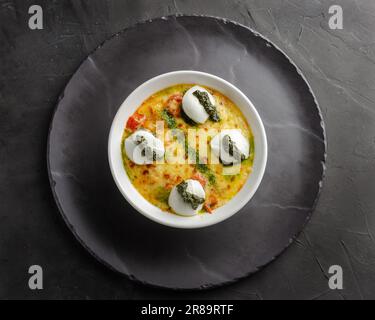 Gnocchi de pomme de terre au gratin au fromage mozzarella et sauce pesto. Servi dans des plats noirs de gibier sur fond noir. Banque D'Images