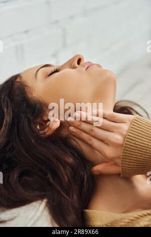 Jeune femme brune en cavalier brun touchant le nœud lymphatique sur le cou et se relaxant pendant l'auto-massage sur le canapé à la maison, rituel d'auto-soins et holistique Banque D'Images