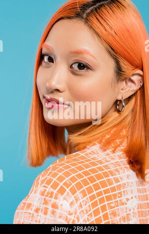 portrait de jeune femme asiatique avec maquillage naturel, brillant à lèvres, boucles d'oreilles et cheveux courts teints regardant l'appareil photo sur fond bleu, jeune, generatio Banque D'Images