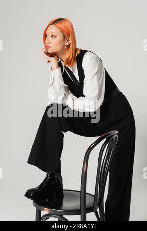 femme asiatique élégante et rêveuse avec des cheveux rouges colorés sur une chaise et regardant loin sur fond gris, chemise blanche, veste et pantalon noirs, affaires Banque D'Images