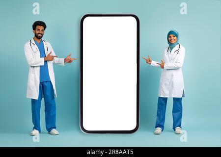 Souriant médecins musulmans en uniforme pointant les doigts sur l'écran de smartphone Big Blank Banque D'Images