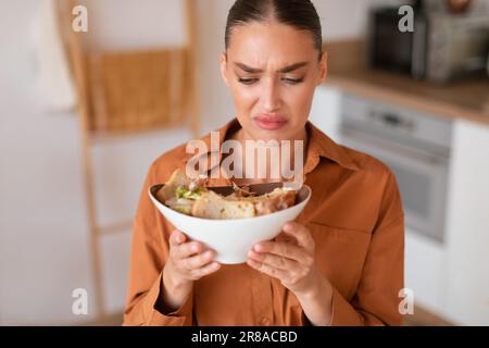 Jeune femme insatisfaite mangeant de la salade césar à la regarder, ne l'aime pas sentir et se sentir dégoût, assis dans la cuisine Banque D'Images