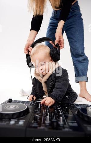 Un enfant mignon dans un sweat à capuche beige et une veste de moto noire drôle se trouve sur le sol avec une console de mixage dj. Une maman pieds nus en Jean bleu met la tête de DJ Banque D'Images