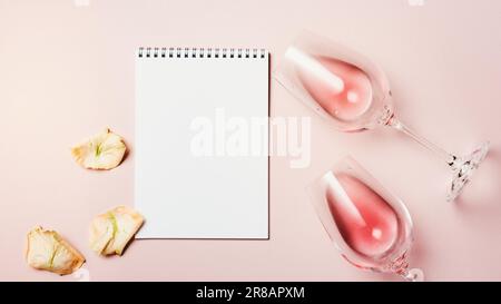 Maquette vierge carnet, deux verres de vin et pétales de rose sur fond rose, vue du dessus. Banque D'Images