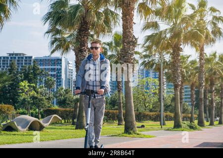 Jeune homme à cheval électrique transport écologique le long de la promenade de la ville avec des palmiers. Homme conduisant e-scooter durable et Banque D'Images