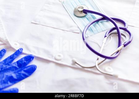 Gros plan d'un stéthoscope et d'un masque chirurgical sur un manteau médical blanc et une partie d'un gant en latex bleu. Banque D'Images