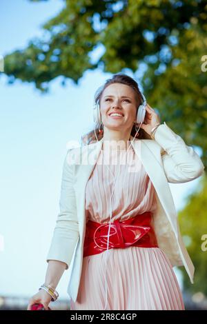 femme élégante et souriante de 40 ans en robe rose et veste blanche en ville avec écouteurs. Banque D'Images