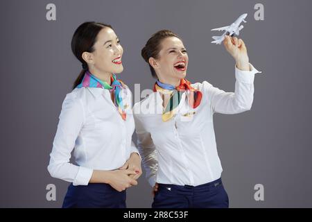 souriante, femme d'hôtesse moderne en jupe bleue, chemise blanche et écharpe avec un petit avion isolé sur fond gris. Banque D'Images