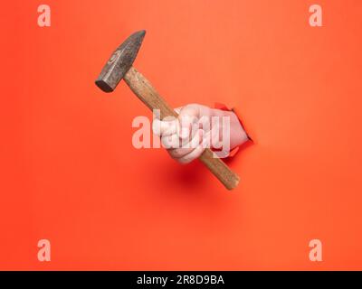 La main d'un homme tient un marteau à travers du papier orange déchiré. Concept, passer à travers le papier orange déchiré. Banque D'Images