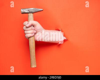 La main d'un homme tient un marteau à travers du papier orange déchiré. Concept, passer à travers le papier orange déchiré. Banque D'Images