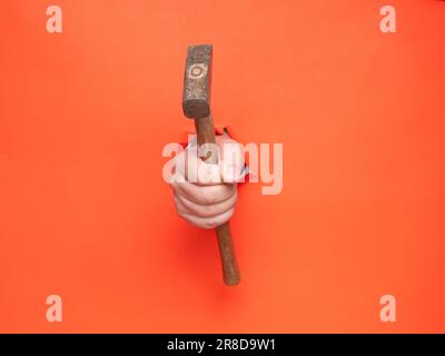 La main d'un homme tient un marteau à travers du papier orange déchiré. Concept, passer à travers le papier orange déchiré. Banque D'Images