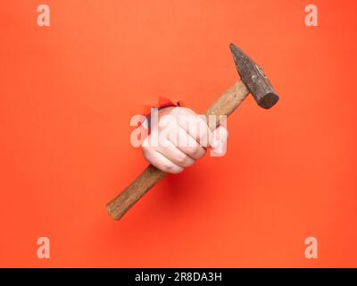 La main d'un homme tient un marteau à travers du papier orange déchiré. Concept, passer à travers le papier orange déchiré. Banque D'Images