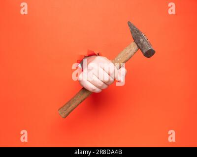 La main d'un homme tient un marteau à travers du papier orange déchiré. Concept, passer à travers le papier orange déchiré. Banque D'Images