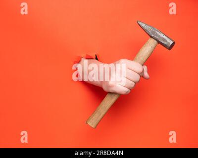 La main d'un homme tient un marteau à travers du papier orange déchiré. Concept, passer à travers le papier orange déchiré. Banque D'Images