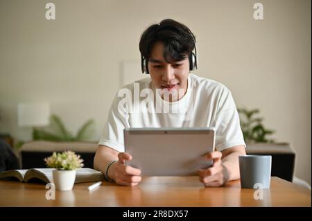 Un jeune homme asiatique heureux vêque de vêtements décontractés porte un casque et aime jouer à des jeux ou regarder des films sur sa tablette numérique à une table dans sa vie Banque D'Images