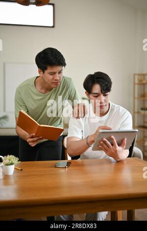 Deux jeunes étudiants asiatiques spécialisés font leurs devoirs à la maison ensemble, à la recherche d'informations dans un livre et sur Internet. Banque D'Images