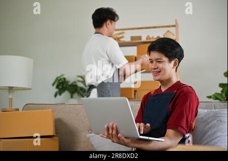 Un jeune homme asiatique heureux est un vendeur en ligne, en vérifiant les commandes en ligne et en mettant à jour de nouveaux produits sur le site de shopping pendant que son petit ami vérifie Banque D'Images