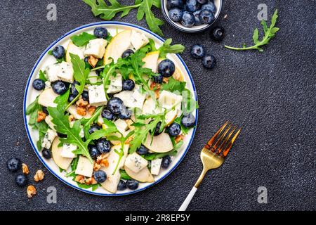 Salade gastronomique aux poires sucrées, aux bleuets, au fromage bleu, à l'arugula et aux noix. Arrière-plan de table noir, vue du dessus Banque D'Images