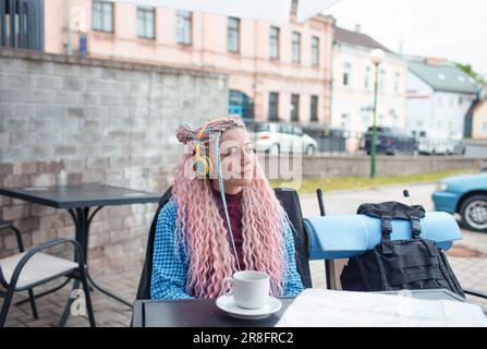 Un jeune touriste séduisant dans une chemise bleue boit du café et écoute de la musique avec des écouteurs. Une fille avec de longs cheveux roses a le déjeuner dans un café. Banque D'Images