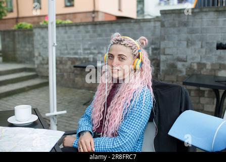 Un jeune touriste séduisant dans une chemise bleue boit du café et écoute de la musique avec des écouteurs. Une fille avec de longs cheveux roses a le déjeuner dans un café. Banque D'Images