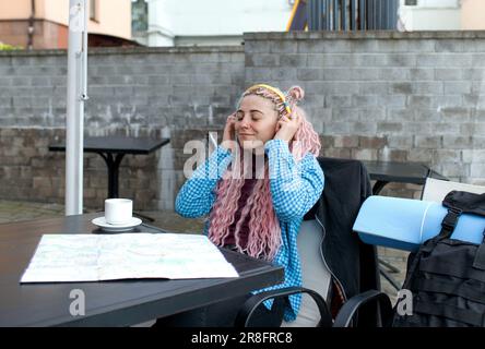 Un jeune touriste séduisant dans une chemise bleue boit du café et écoute de la musique avec des écouteurs. Une fille avec de longs cheveux roses a le déjeuner dans un café. Banque D'Images