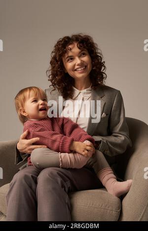 mère moderne qui travaille, concept d'équilibre travail et vie, femme d'affaires heureuse en costume assis avec la petite fille sur un fauteuil, s'engageant avec l'enfant, gr Banque D'Images