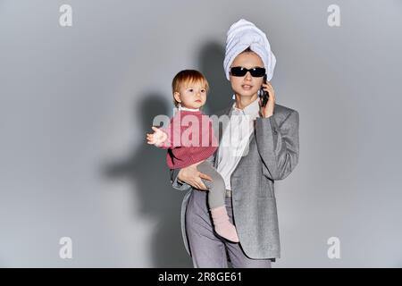 multitâche, femme en lunettes de soleil tenant dans les bras petite fille et debout avec une serviette sur la tête, équilibrant le style de vie, femme d'affaires en tenue formelle Banque D'Images