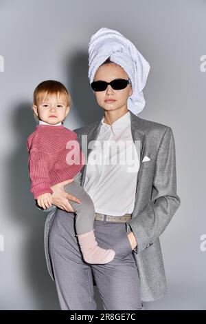 multitâche, femme dans des lunettes de soleil tenant dans les bras petite fille et debout avec une serviette sur la tête, équilibrant entre le travail et la vie, la femme autonome i Banque D'Images