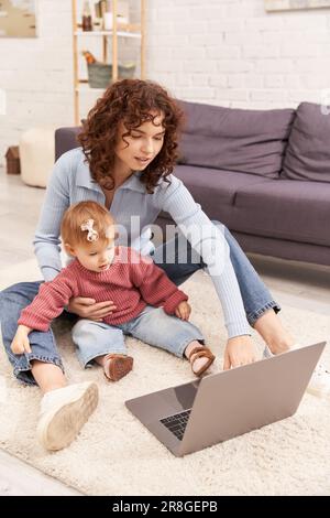 femme multitâche, freelance, parent moderne, s'engager avec l'enfant, équilibrer le travail et la vie, femme curly utilisant un ordinateur portable dans un salon confortable, mode Banque D'Images