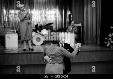 Femme âgée chantant sur scène ancien couple dansant salle de bal 1980s Royaume-Uni. Coventry Working Mens Club, samedi soir la soirée de bingo est terminée et un couple plus âgé bénéficie d'une valse tranquille. Un chanteur local se produit sur scène. Coventry, Angleterre vers 1981. 80S ROYAUME-UNI HOMER SYKES Banque D'Images