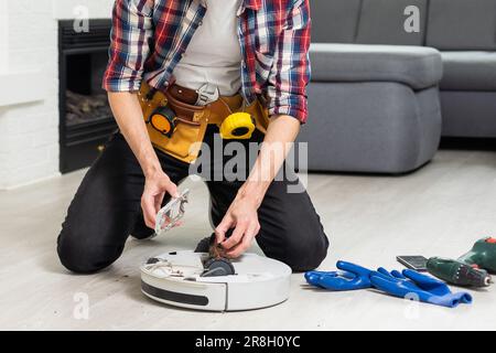 Un jeune homme de race blanche effectue la maintenance d'un robot-aspirateur blanc. Aspirateur moderne sans fil autonome. Nettoyant robot de réparation pour homme à la maison Banque D'Images