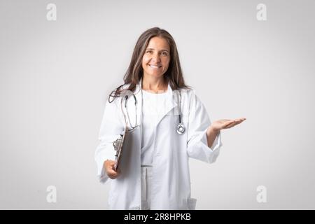 Femme âgée excitée médecin tenant le presse-papiers et montrant quelque chose sur la paume vide, la publicité de poduit ou la clinique Banque D'Images