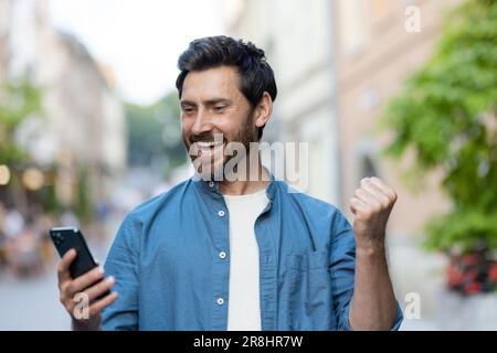 Adulte homme réussi marche dans la ville du soir lors d'un voyage, le gars a reçu une notification avec de bonnes nouvelles par une victoire sur le téléphone, utilise une application en ligne sur un smartphone. Banque D'Images