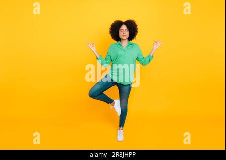 Photo pleine longueur d'une belle et calme souriante femme curly afro-américaine ou brésilienne méditant tout en se tenant debout pose d'arbre avec des yeux fermés sur un fond jaune isolé. Santé mentale, harmonie Banque D'Images