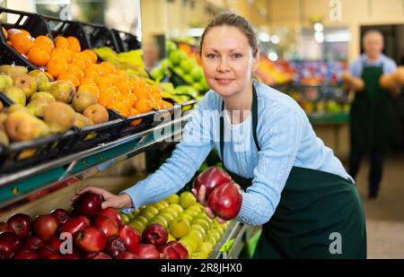 Une femme sympathique d'âge moyen vend des pommes rouges en tablier au supermarché Banque D'Images