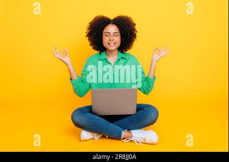 Méditation, tranquillité. Photo en longueur d'une femme afro-américaine ou brésilienne calme et détendue, assise sur un fond jaune isolé avec un ordinateur portable, méditer avec les yeux fermés, prendre soin de la santé mentale Banque D'Images