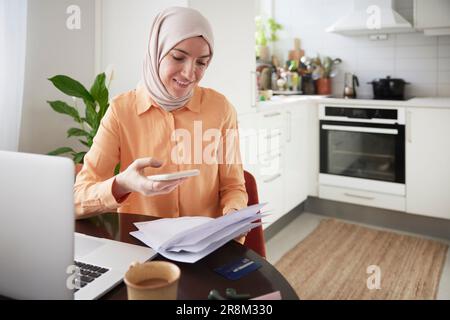 Femme souriante avec hijab utilisant le téléphone cellulaire et de scanner des factures Banque D'Images