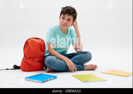 Authentique adolescent d'école assis près de sac à dos orange et des manuels scolaires, raisonnement sur les problèmes d'école, isolé sur blanc Banque D'Images