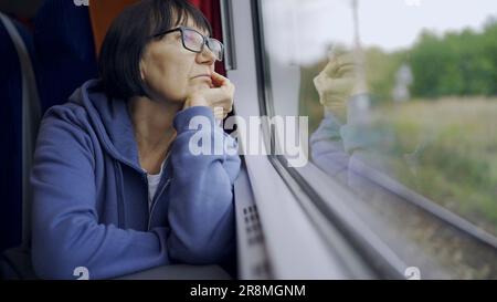 Une femme âgée en verre voyage en train et regarde la fenêtre reflétée dans le verre Banque D'Images