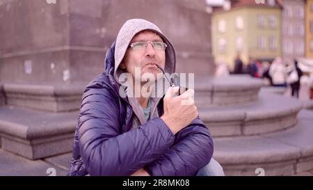 Portrait d'un homme adulte avec des lunettes assis dans la capuche sur la place et fumant un pipe de tabac sur la place du Palais, dans la vieille ville de Varsovie Banque D'Images