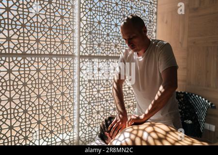 Vue latérale d'un masseur masculin concentré cou massant d'un jeune homme sportif allongé sur une table de massage au soleil et dans de belles ombres Banque D'Images